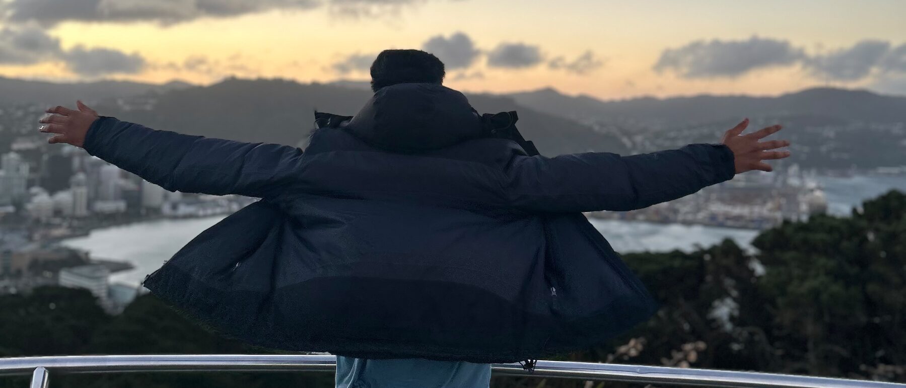 Murali Anand standing arms out at the Mount Victoria lookout above Wellington harbour at sunset